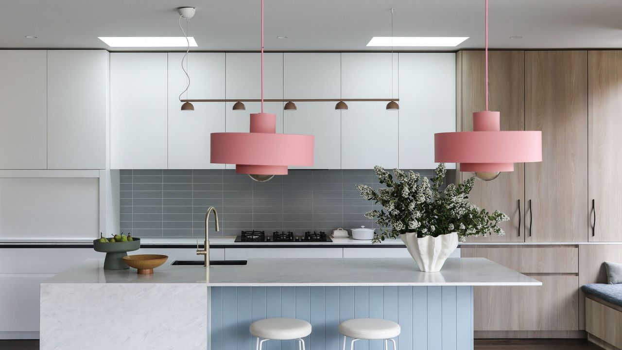 Large pink pendant lights hang above a kitchen island and sink. A chic vase with flowers is placed on top of the island with fruit bowls. Opposite the island are kitchen cabinets and drawers. 