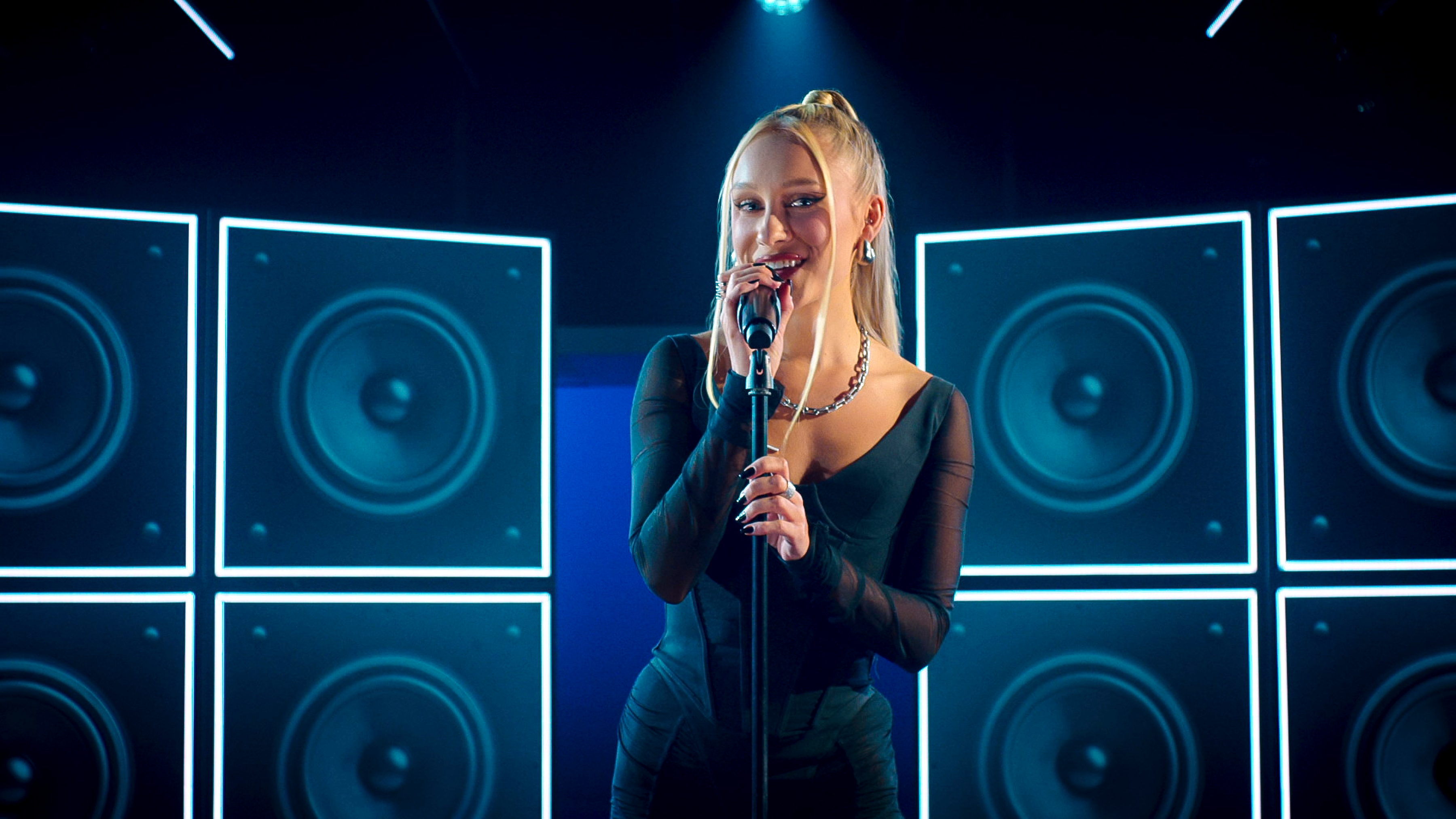 Alison Ogden with her hair in a pony tail, wearing an all black outfit and singing at a microphone in a still from netflix's Building The Band