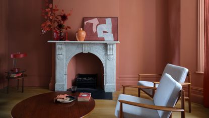 A living room with Farrow & Ball’s Red Earth matt paint on walls and ceiling, with a white marble fireplace and white/teak armchairs