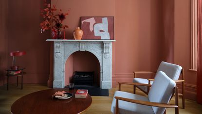 A living room with Farrow &amp; Ball’s Red Earth matt paint on walls and ceiling, with a white marble fireplace and white/teak armchairs