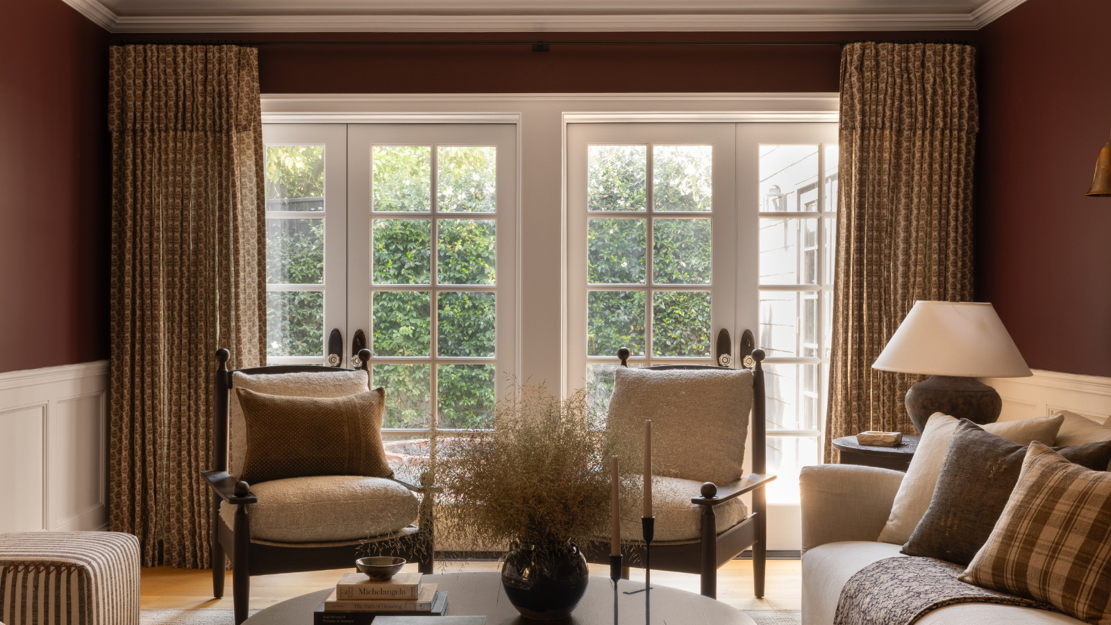 a living room with red walls, a set of two chairs, a coffee table holding books, a vase, and candle holders, and double french doors to the outdoor area framed with floor length curtains