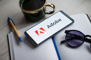 Adobe logo displayed on a smartphone sitting on a desk with coffee mug and notepad.