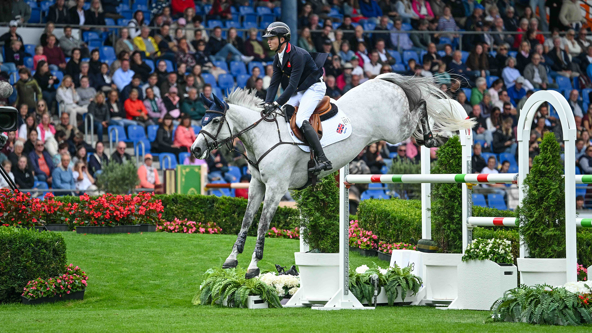 Harry Charles and his grey mare clear a large oxer of mainly white poles, with just a small green and red stripe at each end, during the speed leg of the showjumping World Champions in Aachen