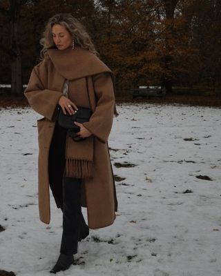 Woman wearing tan scarf coat, walking in snow.