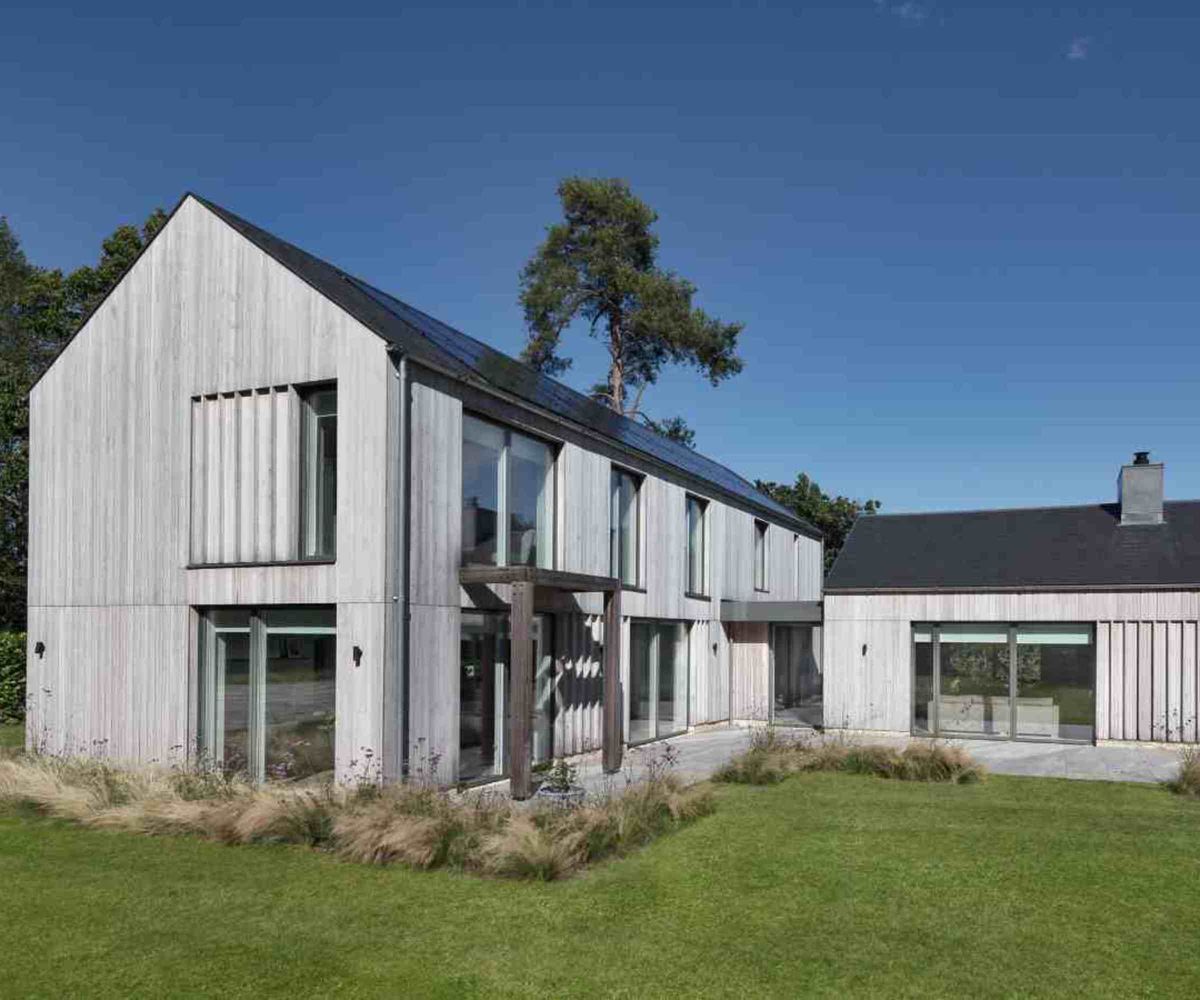 L-shaped timber-clad self build home, with lots of windows and sliding doors, from VELFAC. The house is surrounded by a neat lawn and trees in the background.
