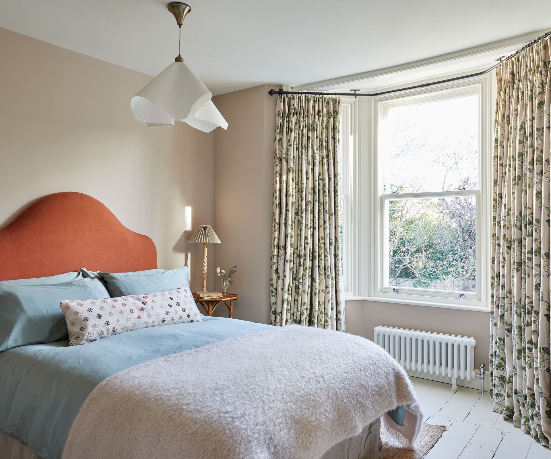 Bedroom with pale pink walls, floral curtains, orange headboard, blue bedding and pink throw