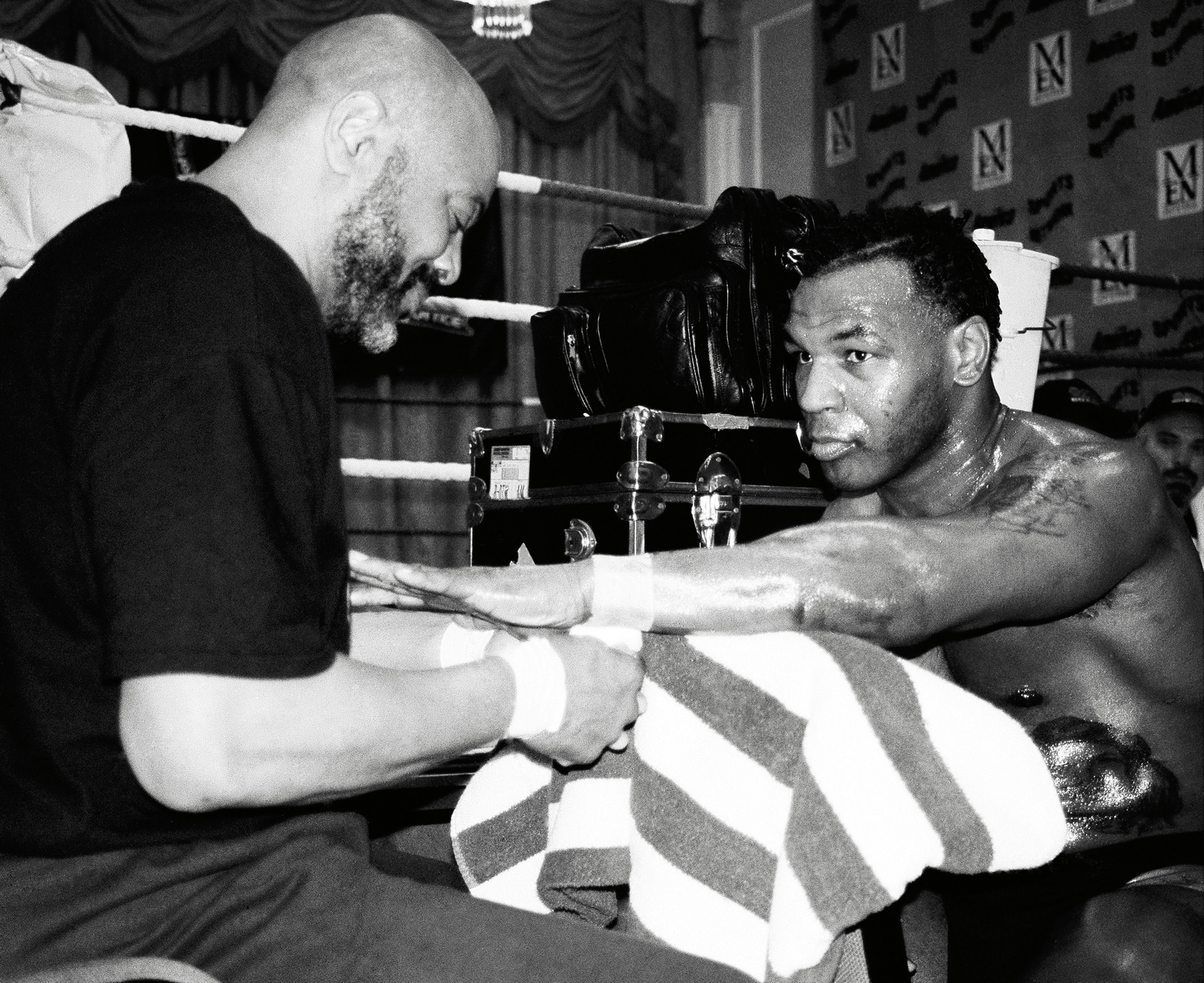 Mike Tyson stares intently at the camera while a cornerman wraps his hands in the corner of a boxing ring, his gloves resting on a case behind him.