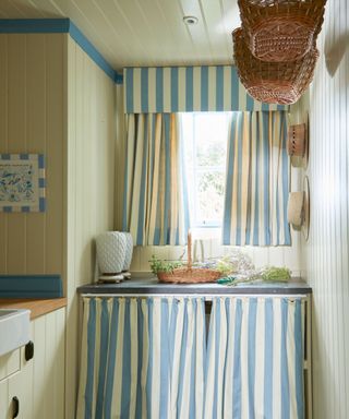Small kitchen with blue and white striped curtains used on the windows and cupboard, blue painted ceiling trim, yellow painted paneled walls, woven baskets hanging from ceiling