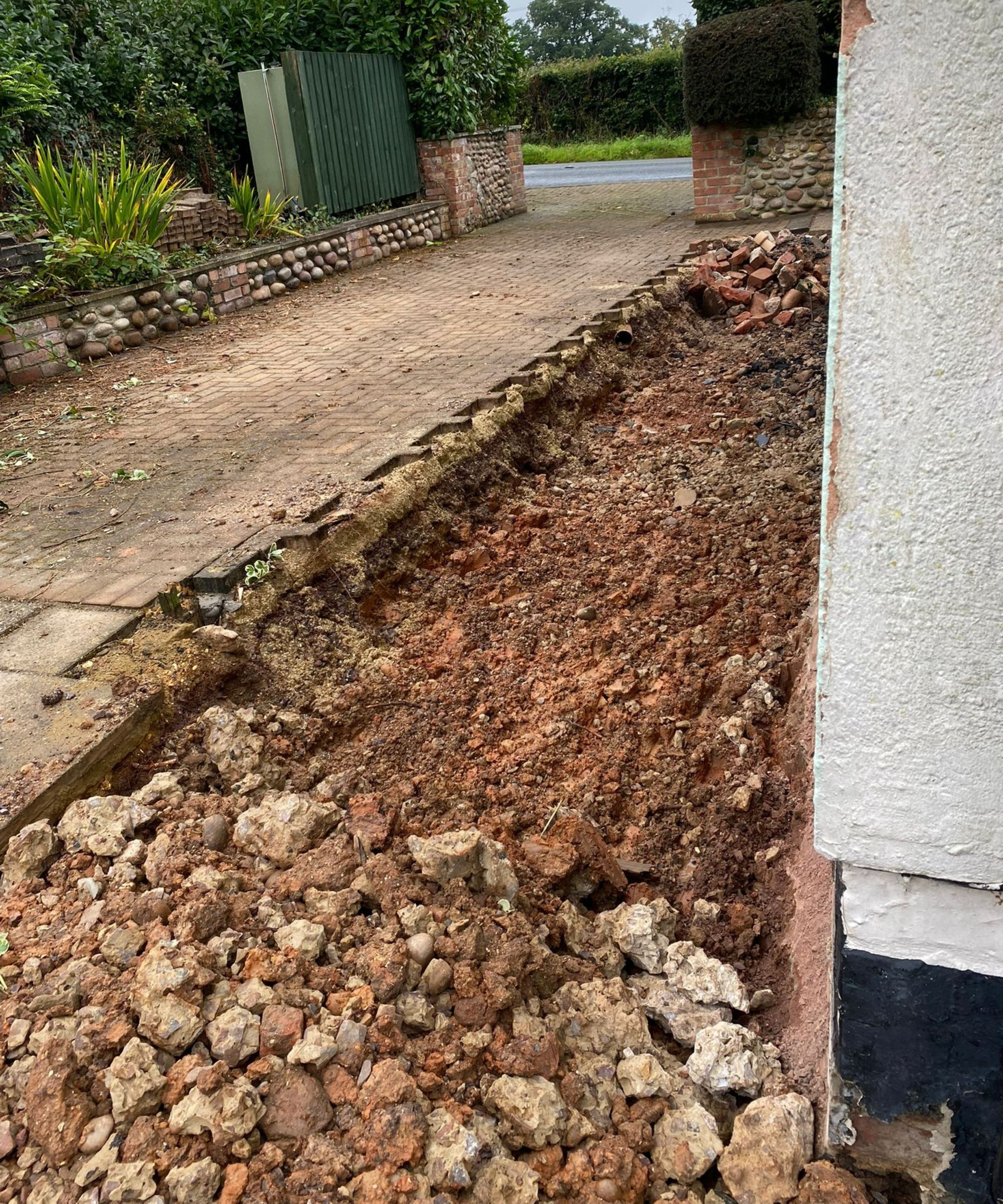 A driveway excavation showing a long trench dug beside a house wall. Exposed soil, rubble and broken bricks line the edge where paving has been removed. Surrounding greenery and a brick boundary wall sit beyond the disrupted groundwork