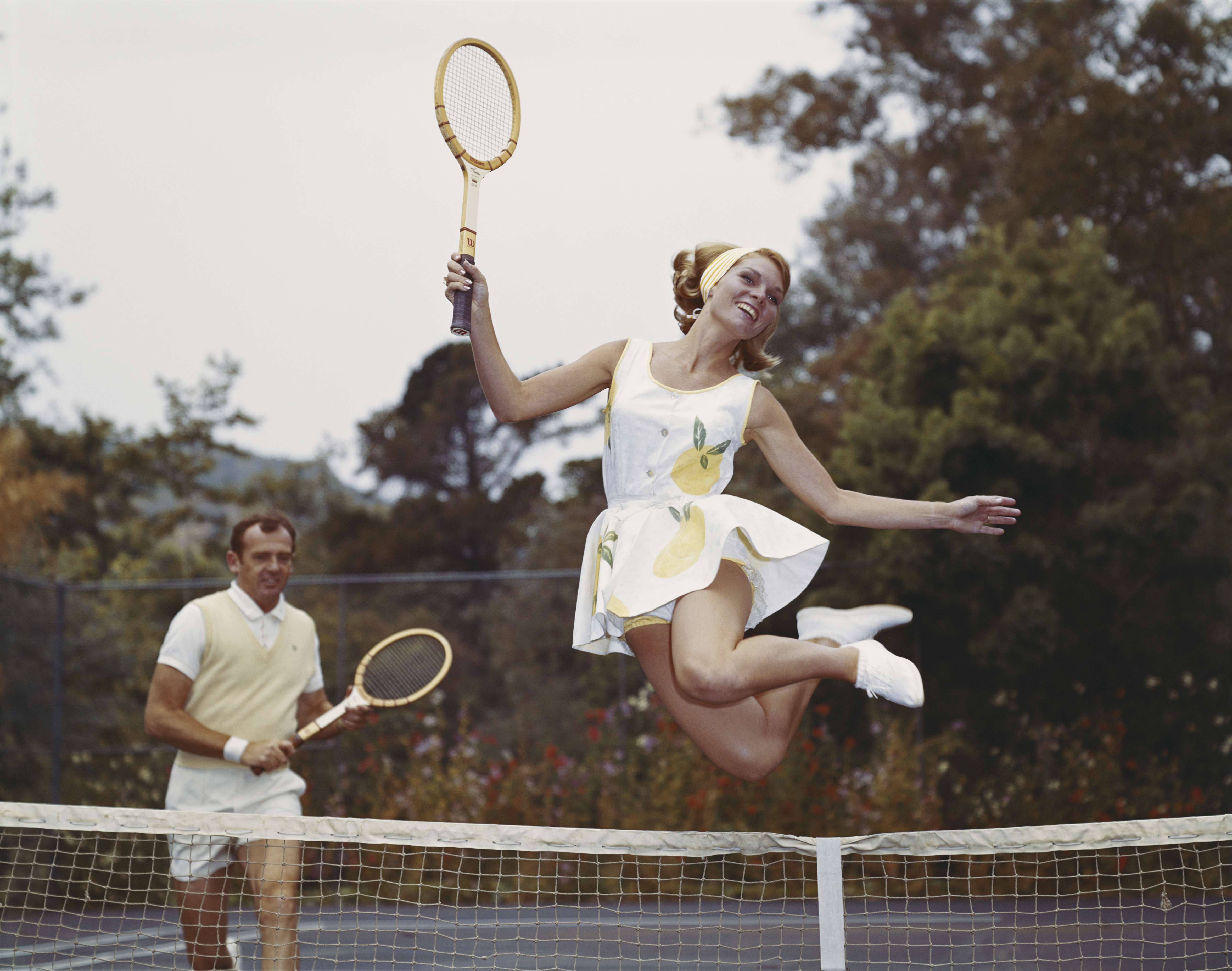 Woman leaps over a tennis court holding a wooden tennis racket in one hand. A man with a matching tennis racket runs behind her