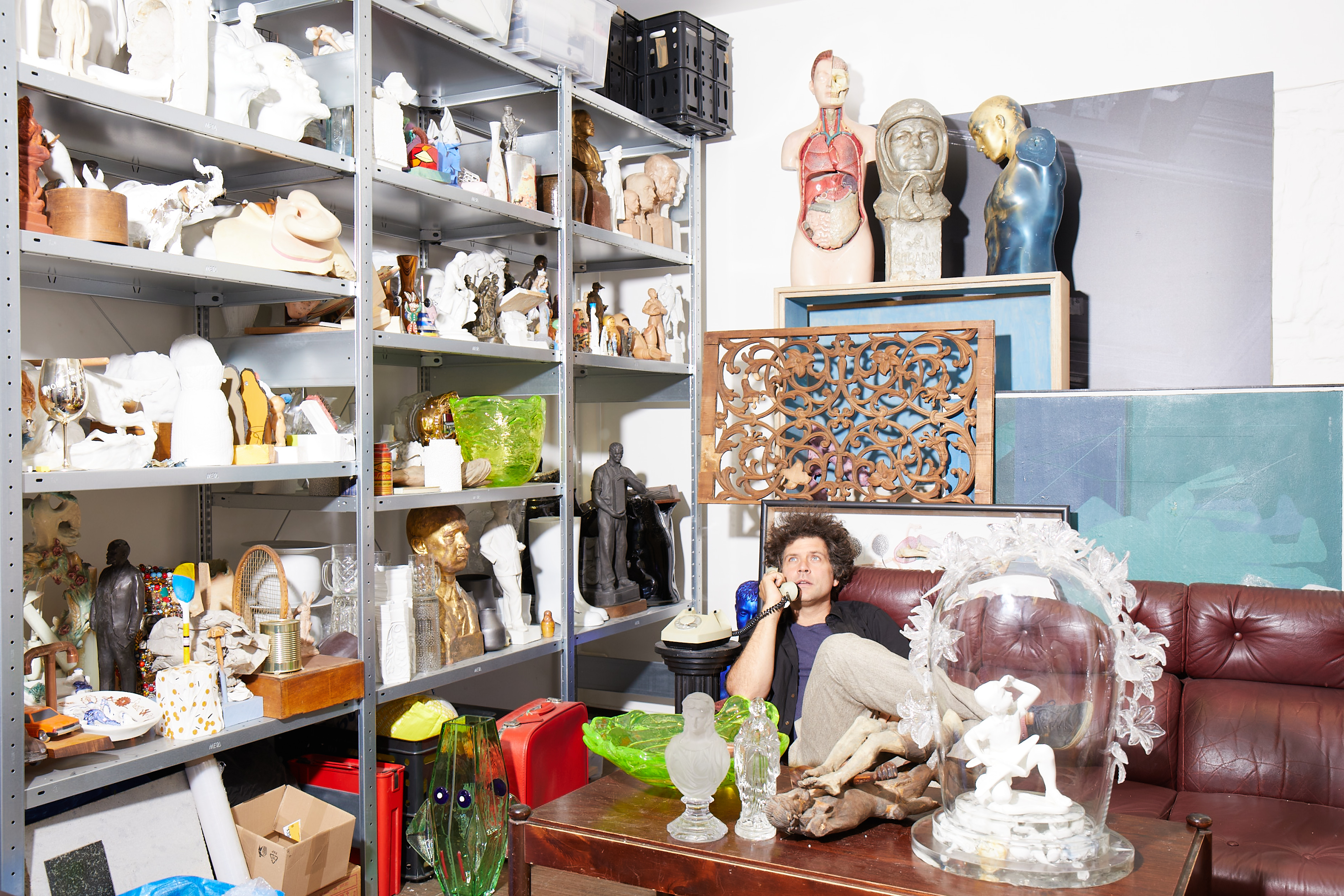 A young man with curly brown hair, dressed in a black blazer with beige trousers and a blue T-shirt underneath, holds a telephone while staring to the ceiling from a leather brown couch, surrounded by quirky creations in a brightly lit artist studio.