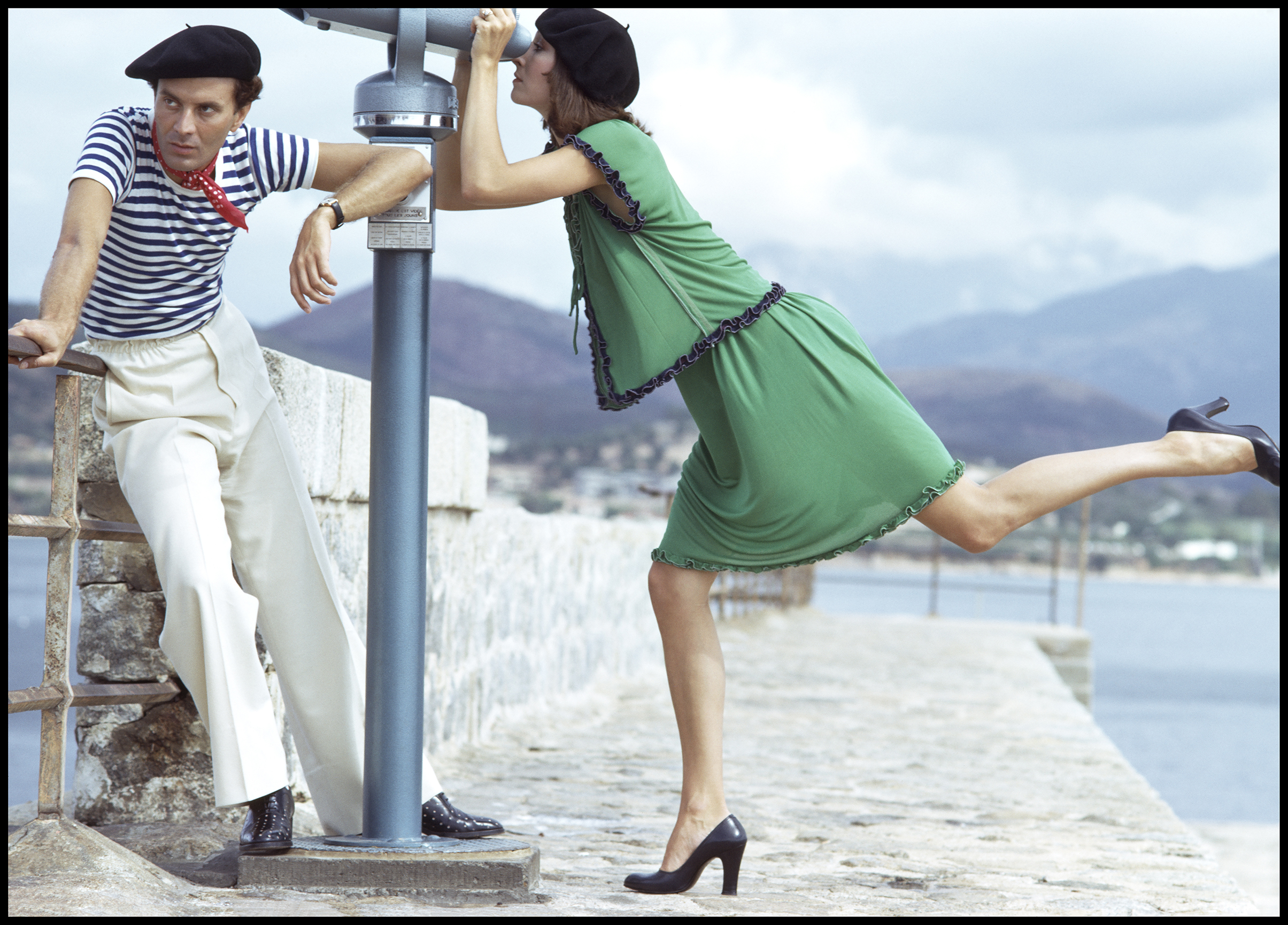 A fashionable couple poses near the water; one leans on a viewing telescope while the other strikes a playful pose in a green dress