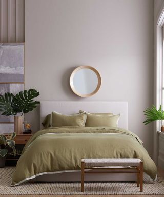 Olive green linen sheets on a cream bed base against a cream wall; to the left, houseplants on a wooden nightstand; hanging above the bed, a circular mirror.