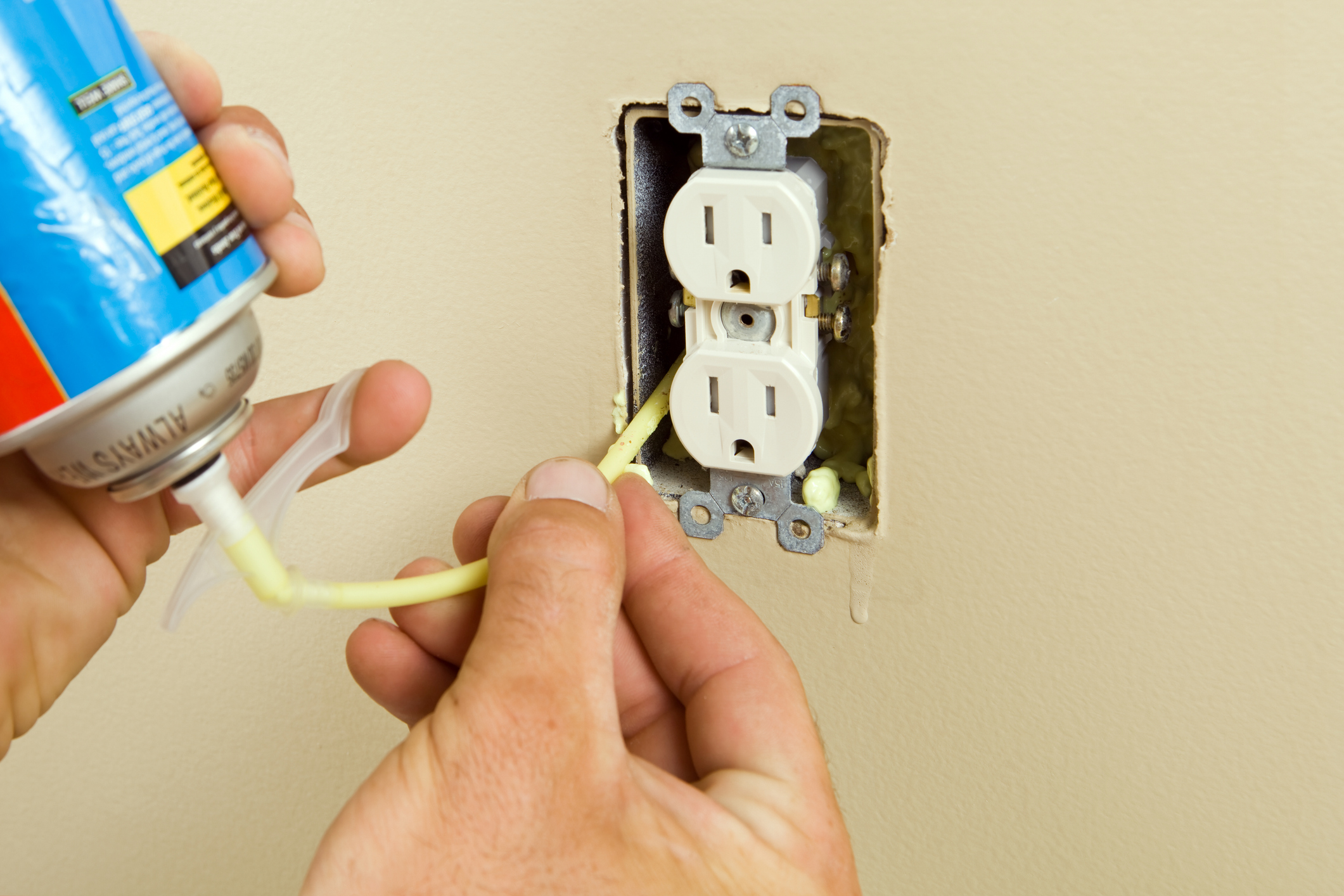 A hands holds a can of expandable foam insulation to insert it into the back of a wall outlet.