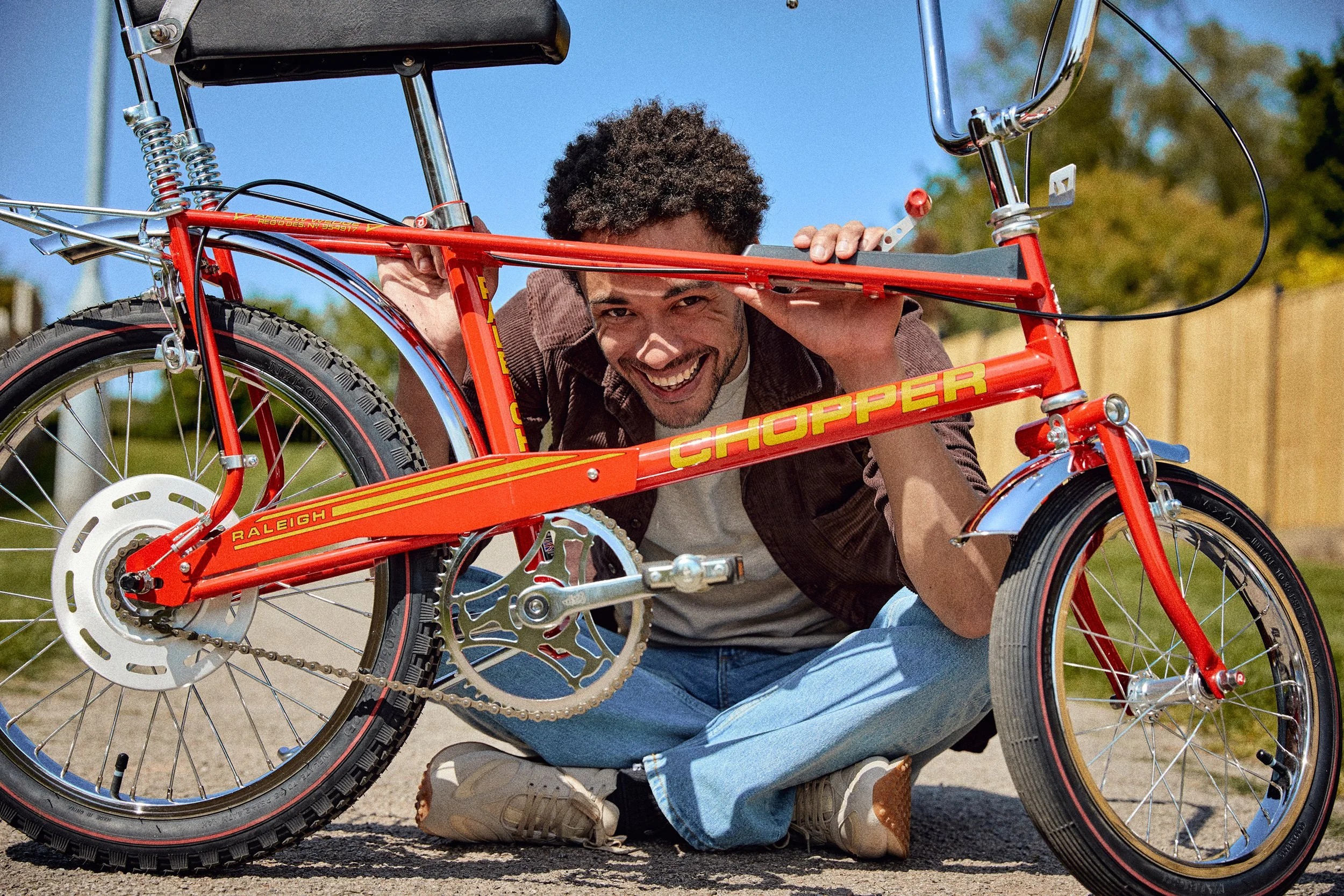 A smiling man sits on the ground behind a bright red Raleigh Chopper bicycle, holding its frame.