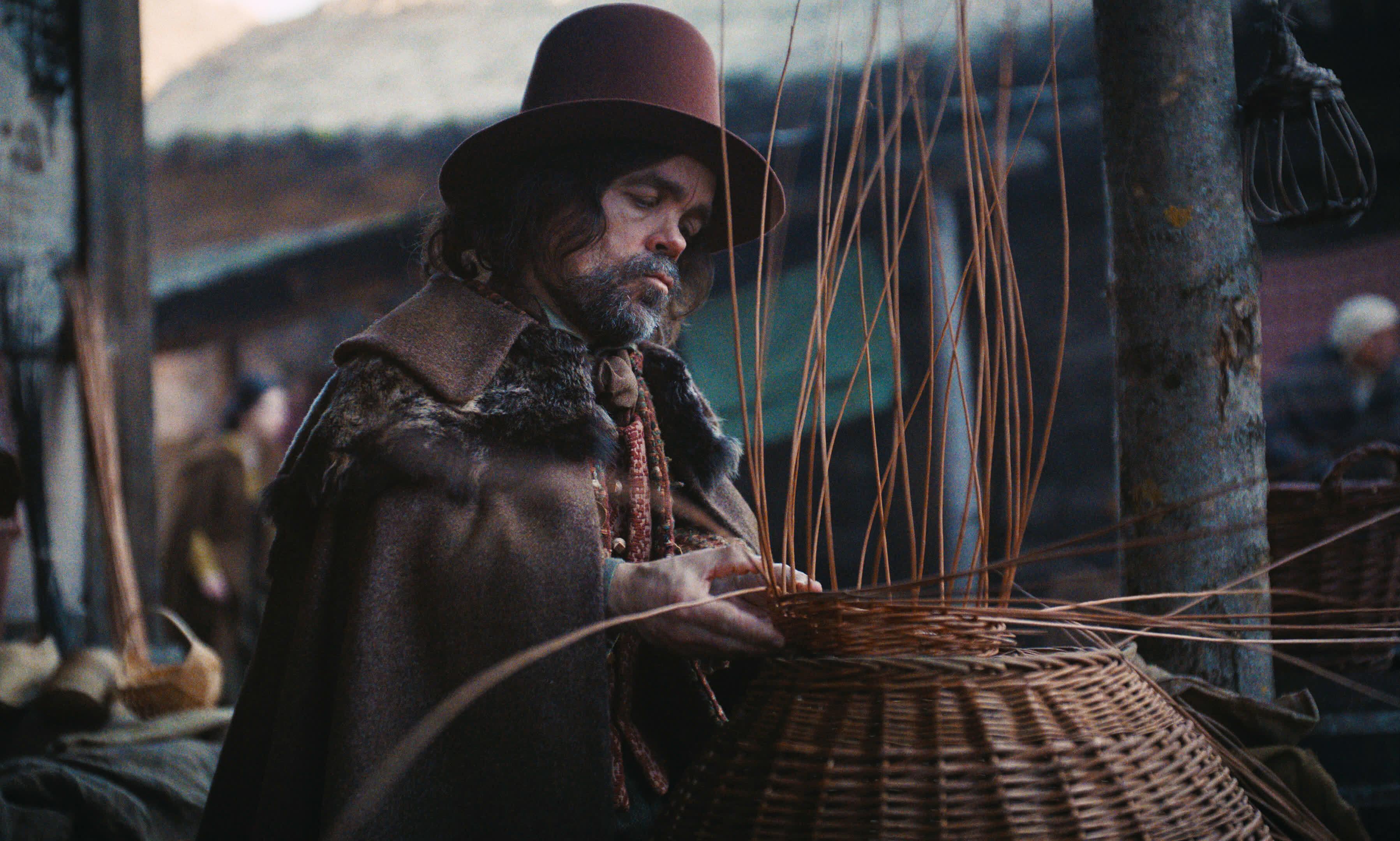 A craftsman, dressed in an old-style brown coat with matching hat, weaves a basket from natural fibers under a village wood-clad patio.