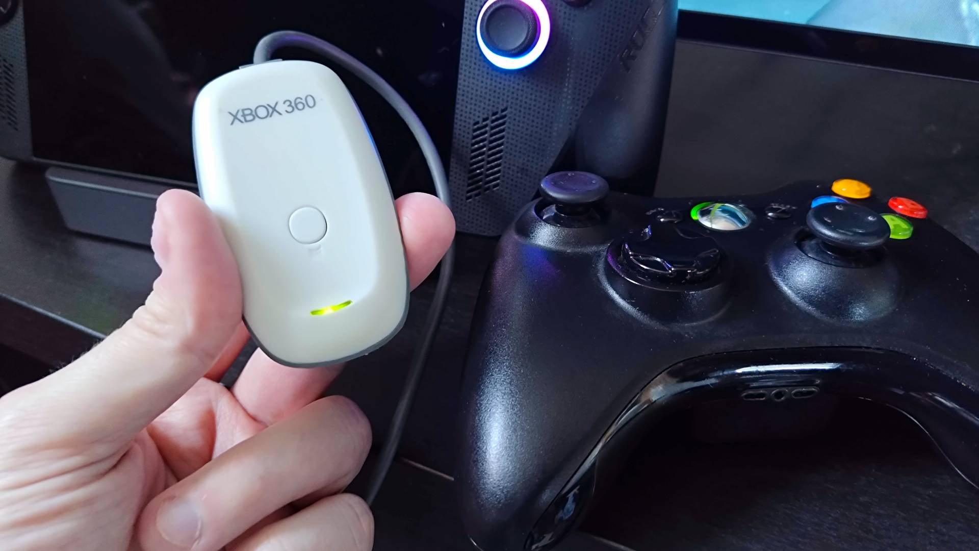 Hand holding Xbox 360 Wireless Controller Adapter for PC next to ROG Xbox Ally X on Baseus Steam Deck dock and black controller.