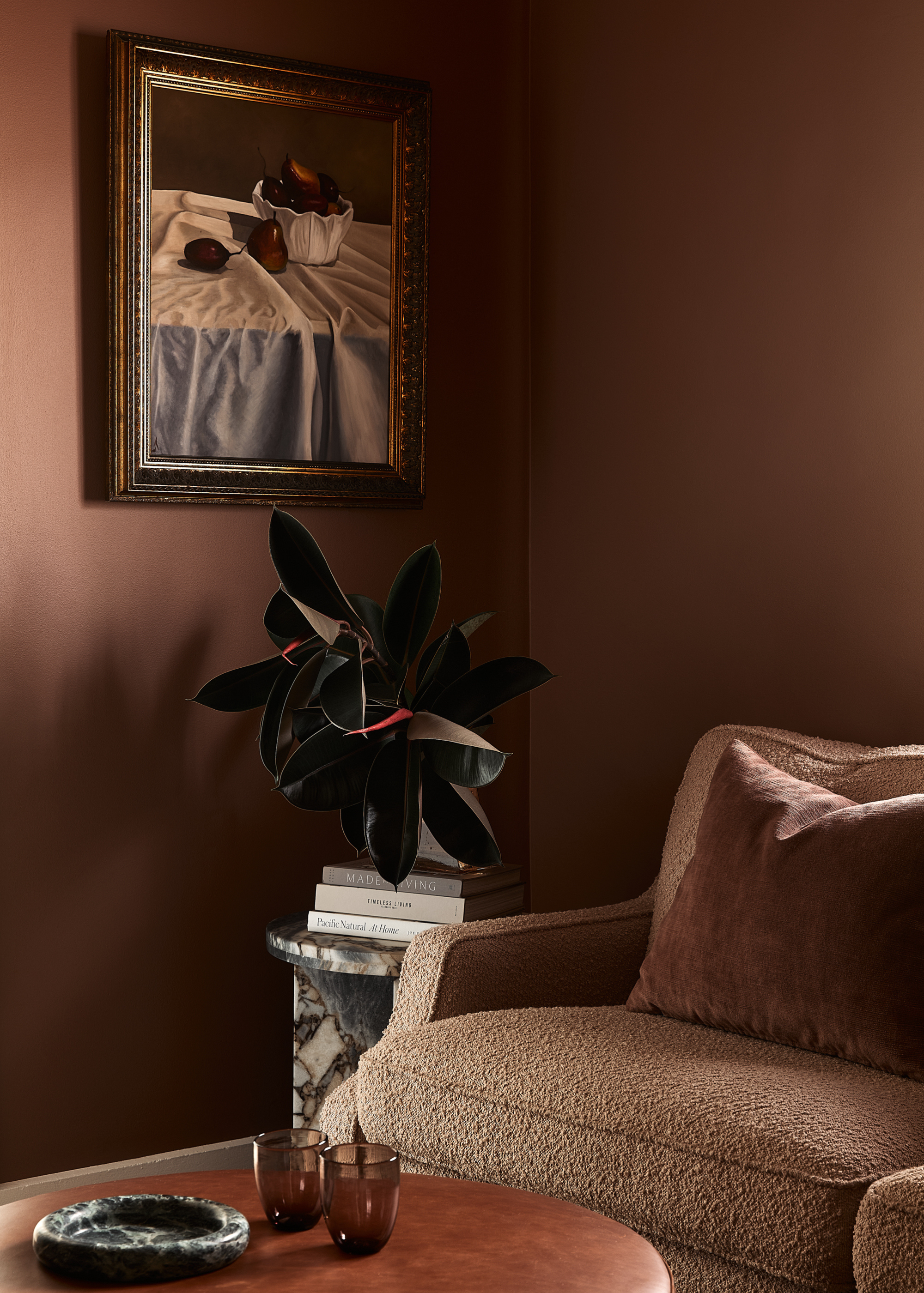 A living room corner with a framed painting of fruit, a potted plant on a marble plinth with a stack of books, by a fuzzy brown couch and a round coffee table with a pair of amber glasses and a marble catch-all
