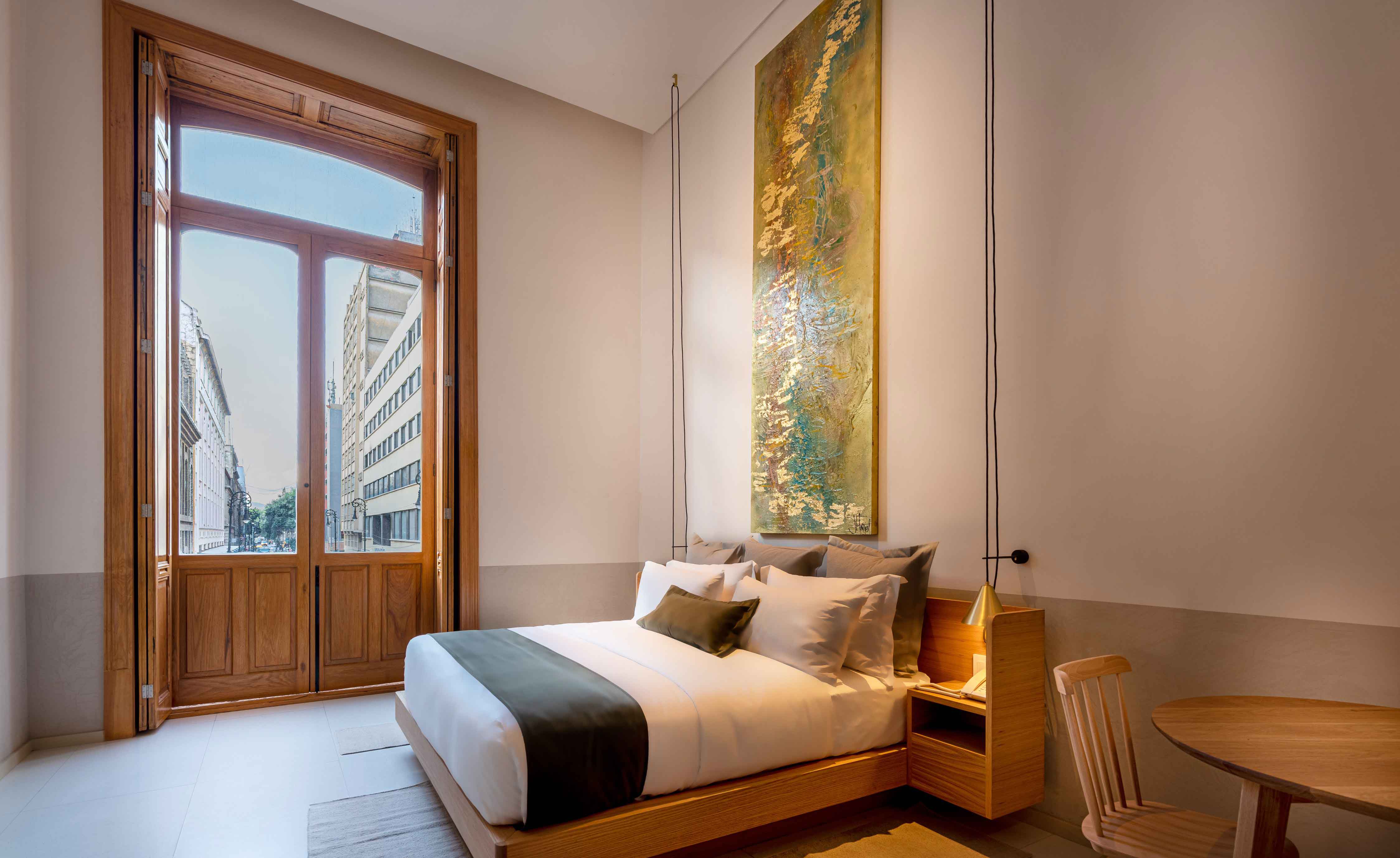 Bedroom with doors onto balony overlooking street, low hanging light features with wooden side tables, chairs with table and bed base