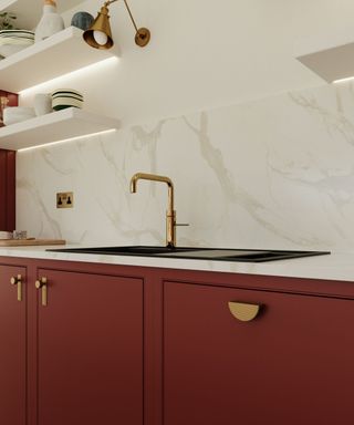 A contemporary kitchen sink with a matte black basin and a gold single-handle tap. The backdrop features a marble-effect backsplash and open shelving with decorative items, complemented by deep red cabinetry with gold hardware and a wall-mounted gold light.