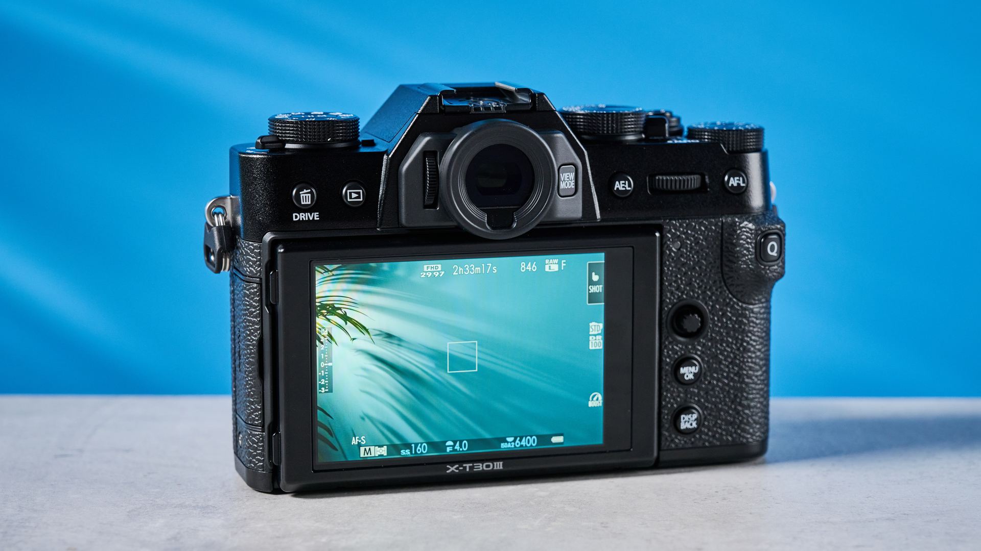 The Fujifilm X-T30 III with its rear display showing, the camera is on a stone surface with a blue background