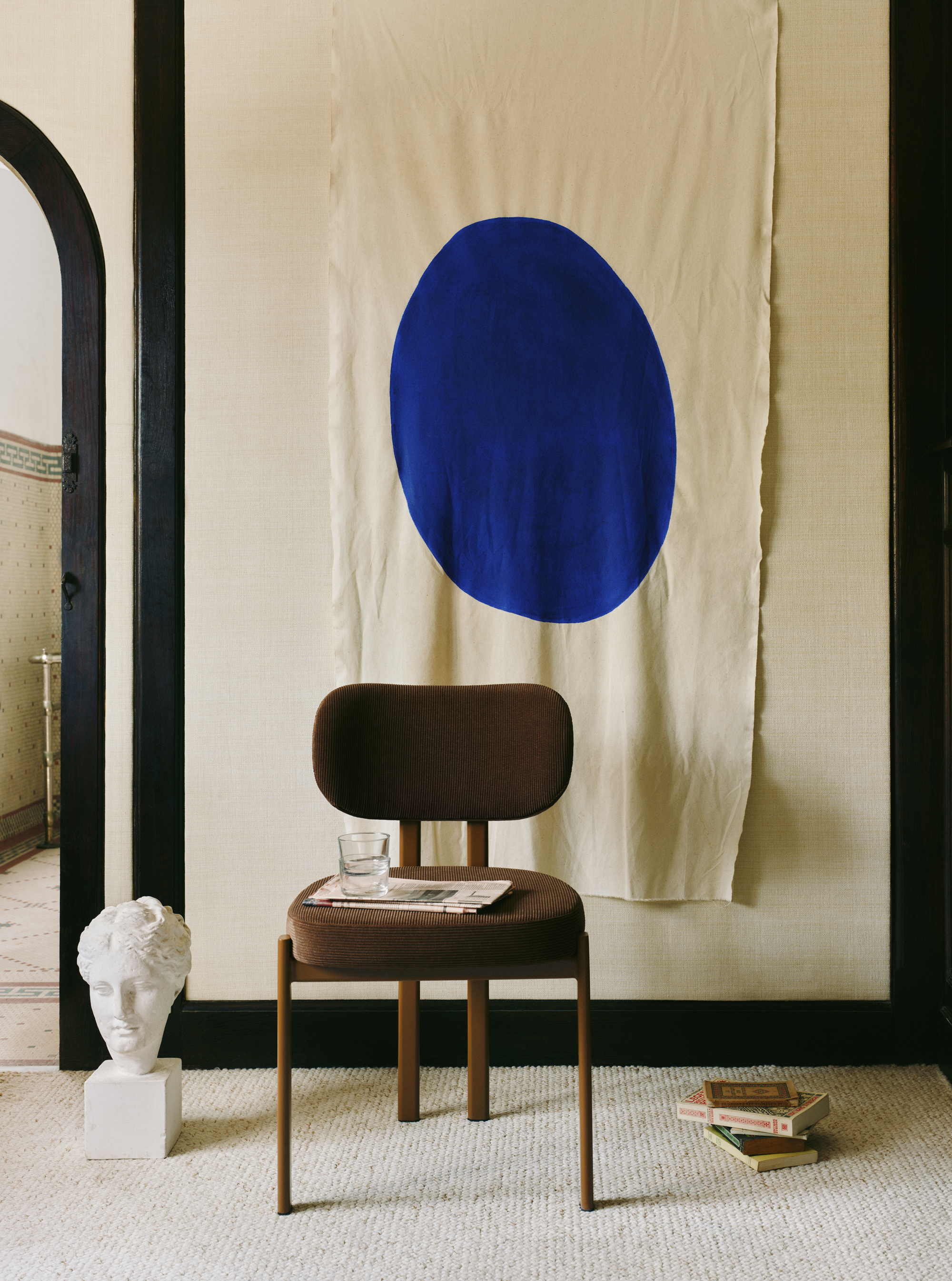 a brown chair with a newspaper and glass styled on the seat, a head bust on the ground and a stack of books, and a cloth with a large blue dot hung on the wall behind