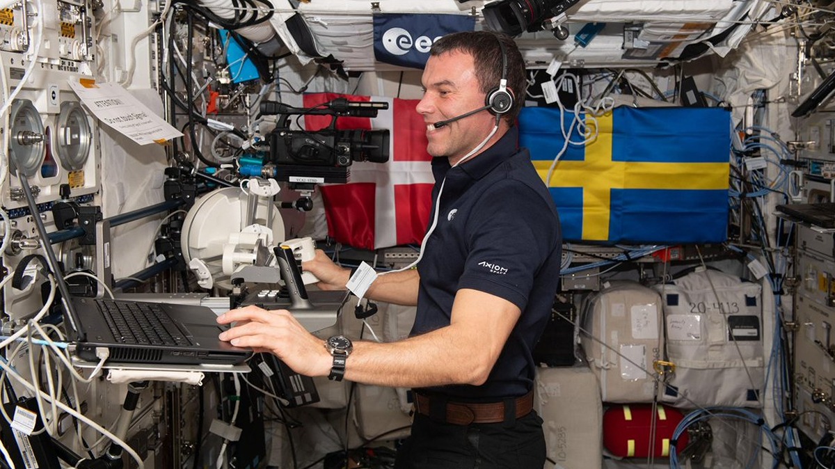 an astronaut at a laptop in the space station backdropped by flags