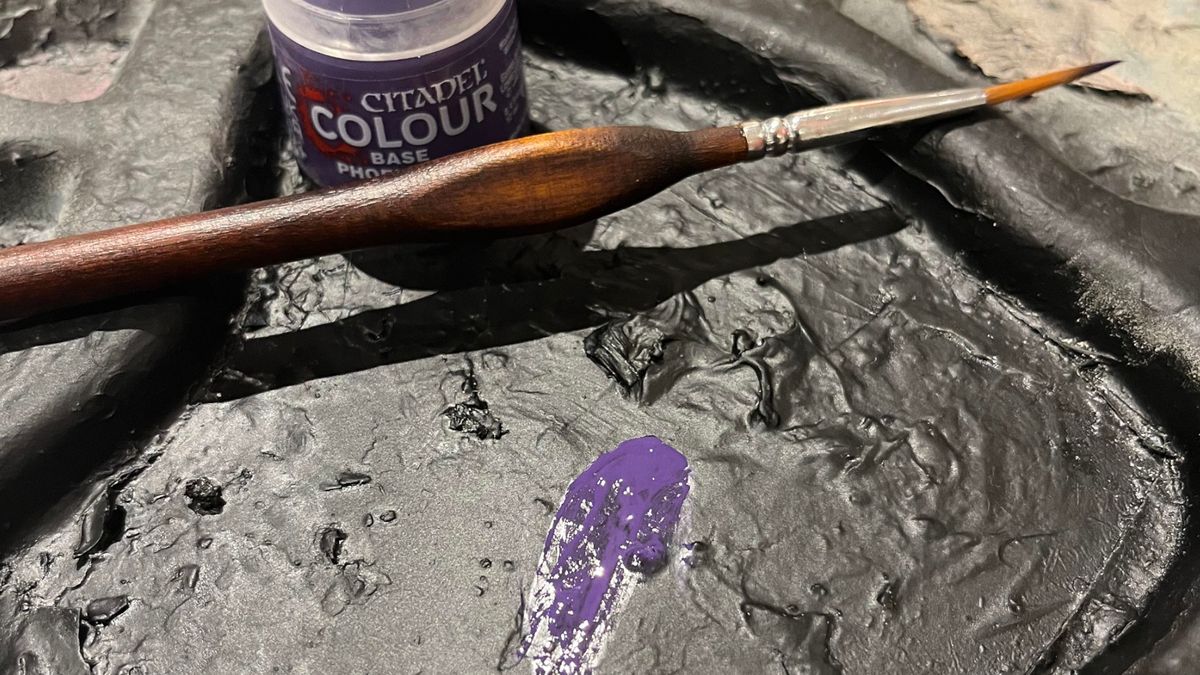 A brush and paint pot sit on a palette covered in black, with purple paint on top