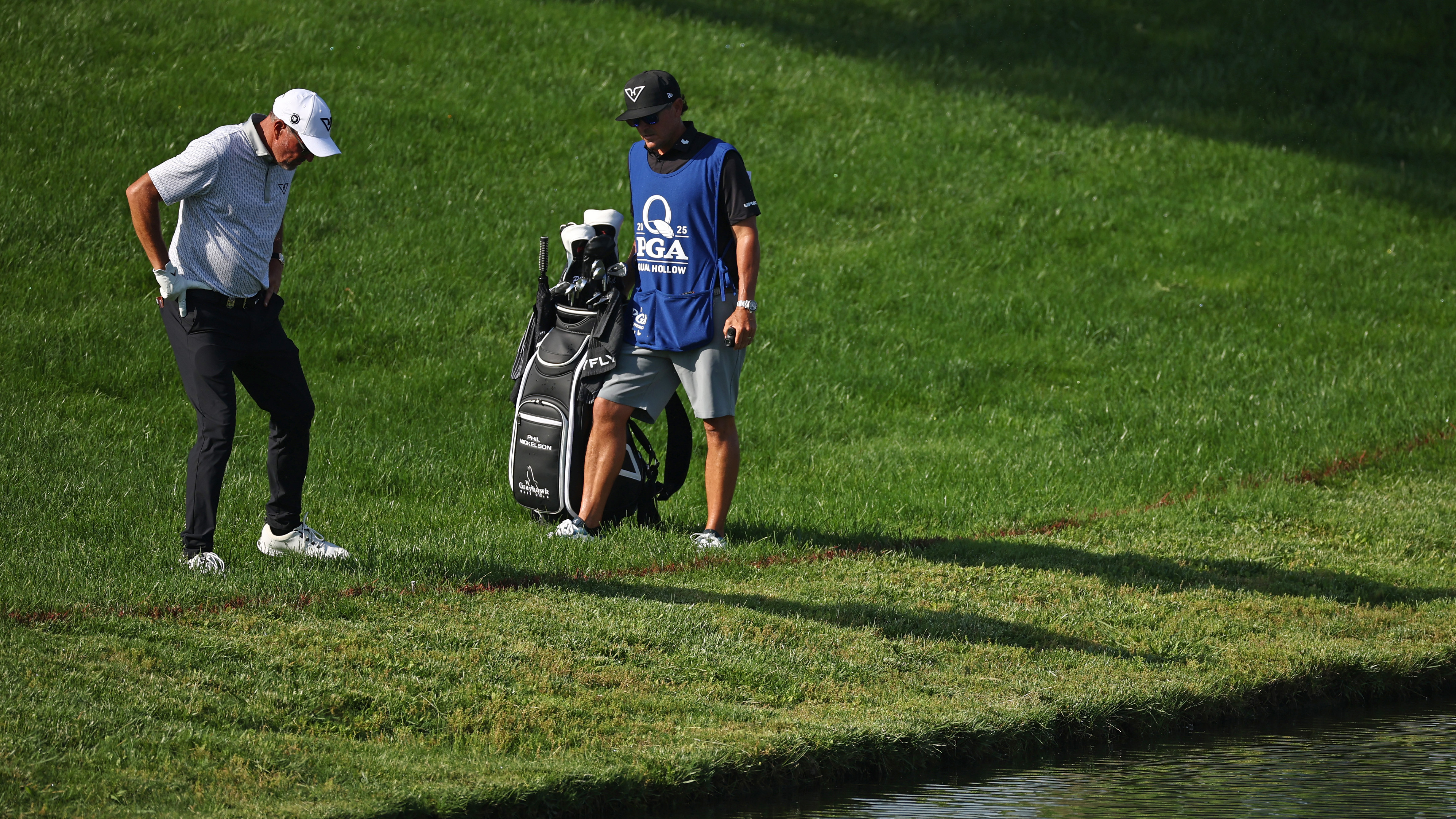 Phil Mickelson and his caddie look down at the rough near water at Quail Hollow during the 2025 PGA Championship