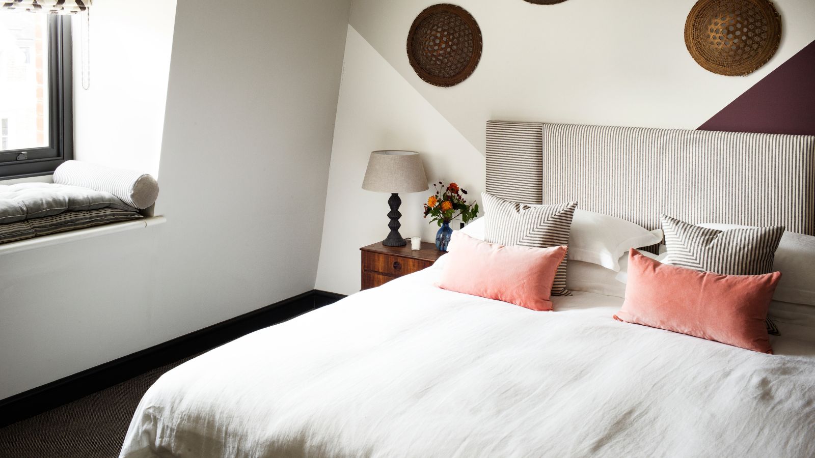 A white bedroom with a large double bed with striped headboard, white bedding, and matching striped and pink cushions. To the left of the bed is a wooden bedside table with a lamp on top, and a large window filling the room with natural light.