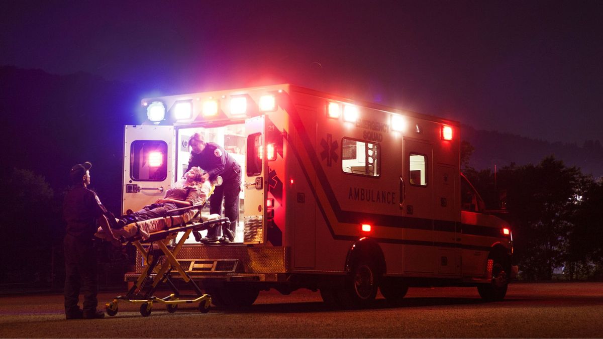 older white woman with grey hair being loaded into ambulance by two paramedics, at night as the ambulance lights flash