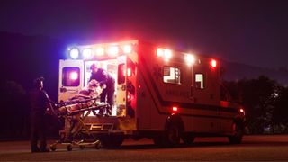 older white woman with grey hair being loaded into ambulance by two paramedics, at night as the ambulance lights flash