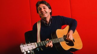 Paul McCartney plays an acoustic guitar against a red background