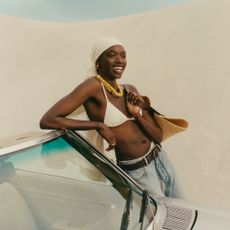 Woman in a white bikini with a white headscarf and jeans sold at Net-a-Porter leaning on a car