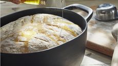 A black cast iron oval pot with a risen bread dough inside, someone pouring oil on top