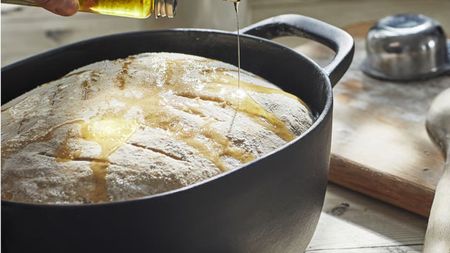 A black cast iron oval pot with a risen bread dough inside, someone pouring oil on top
