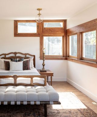 Bedroom with unique square windows with wooden frames, double bed with wooden frame, daybed with striped upholstery at the foot of the bed, wooden flooring