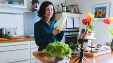 woman standing in a kitchen holding a pointed cabbage and smiling. there are also tulips in shot.