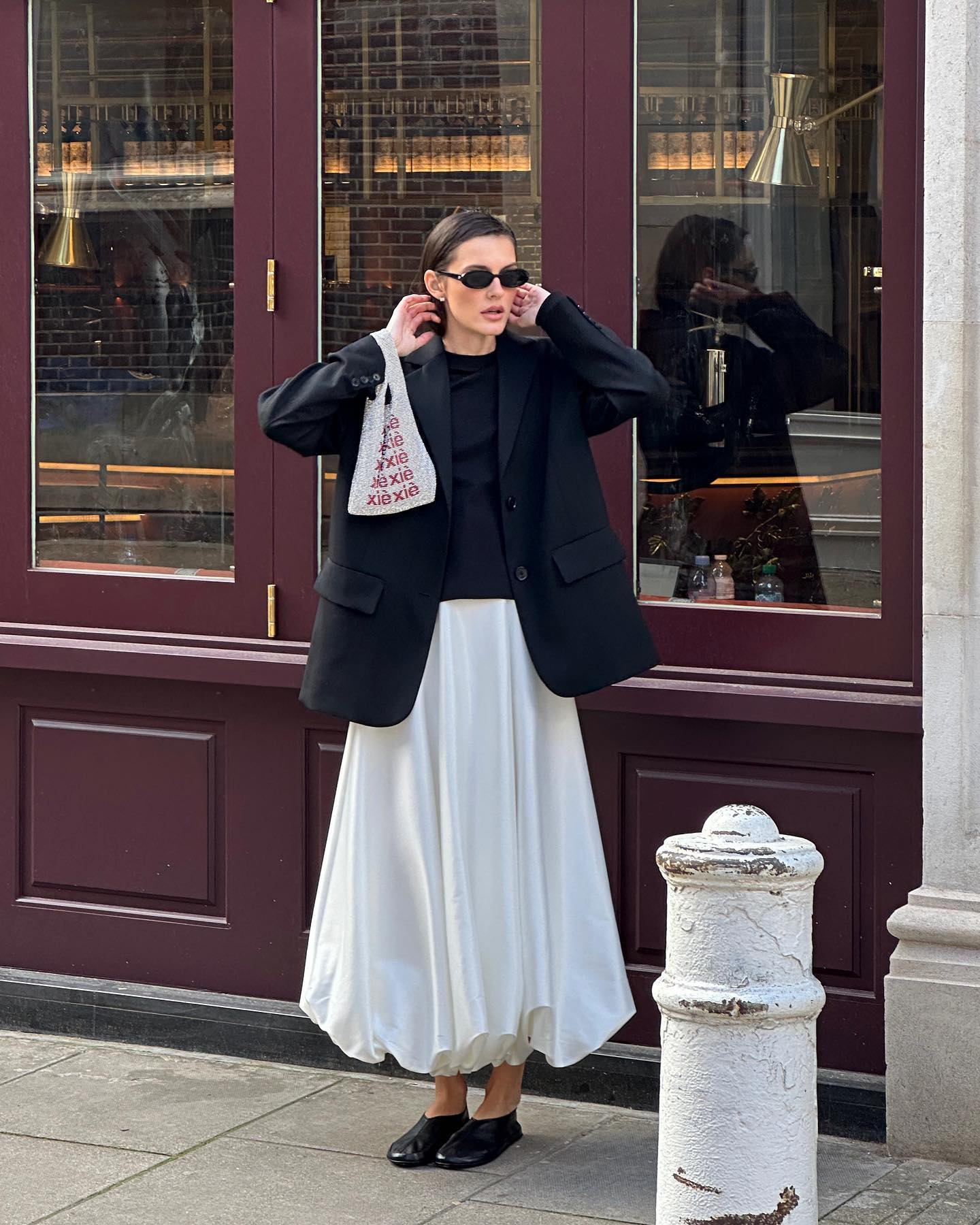 stylish woman poses on the sidewalks of London in black oval sunglasses, a black blazer, black top, white bubble hem skirt and black flats