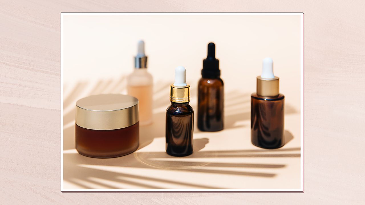 Image showing a collection of unbranded skincare products set on a pale beige surface and in the shadow of a leaf, in a white frame set against a dusky pink watercolour-style background