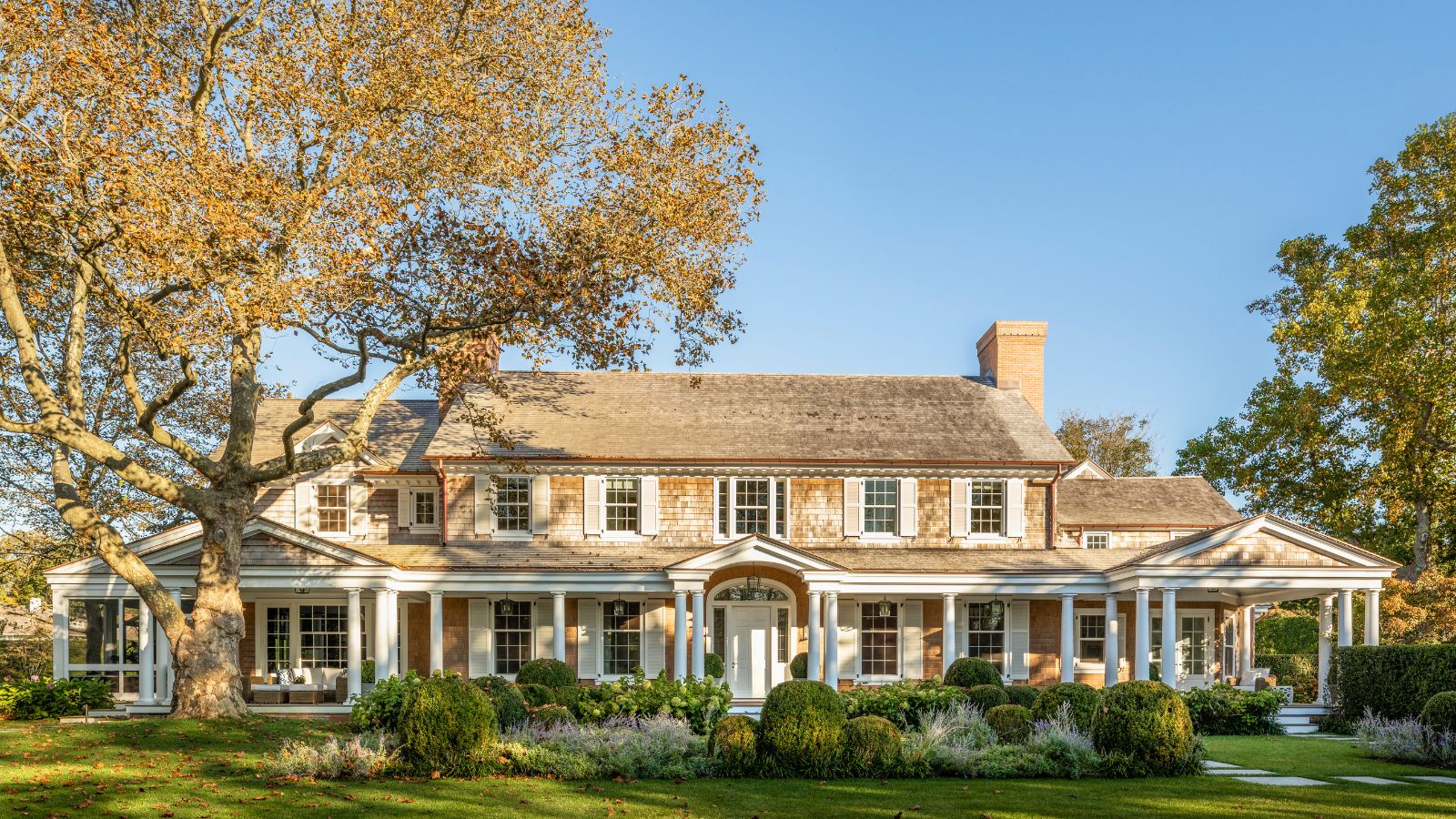 Shingle style home with columns and a long porch, large tree and green lawn