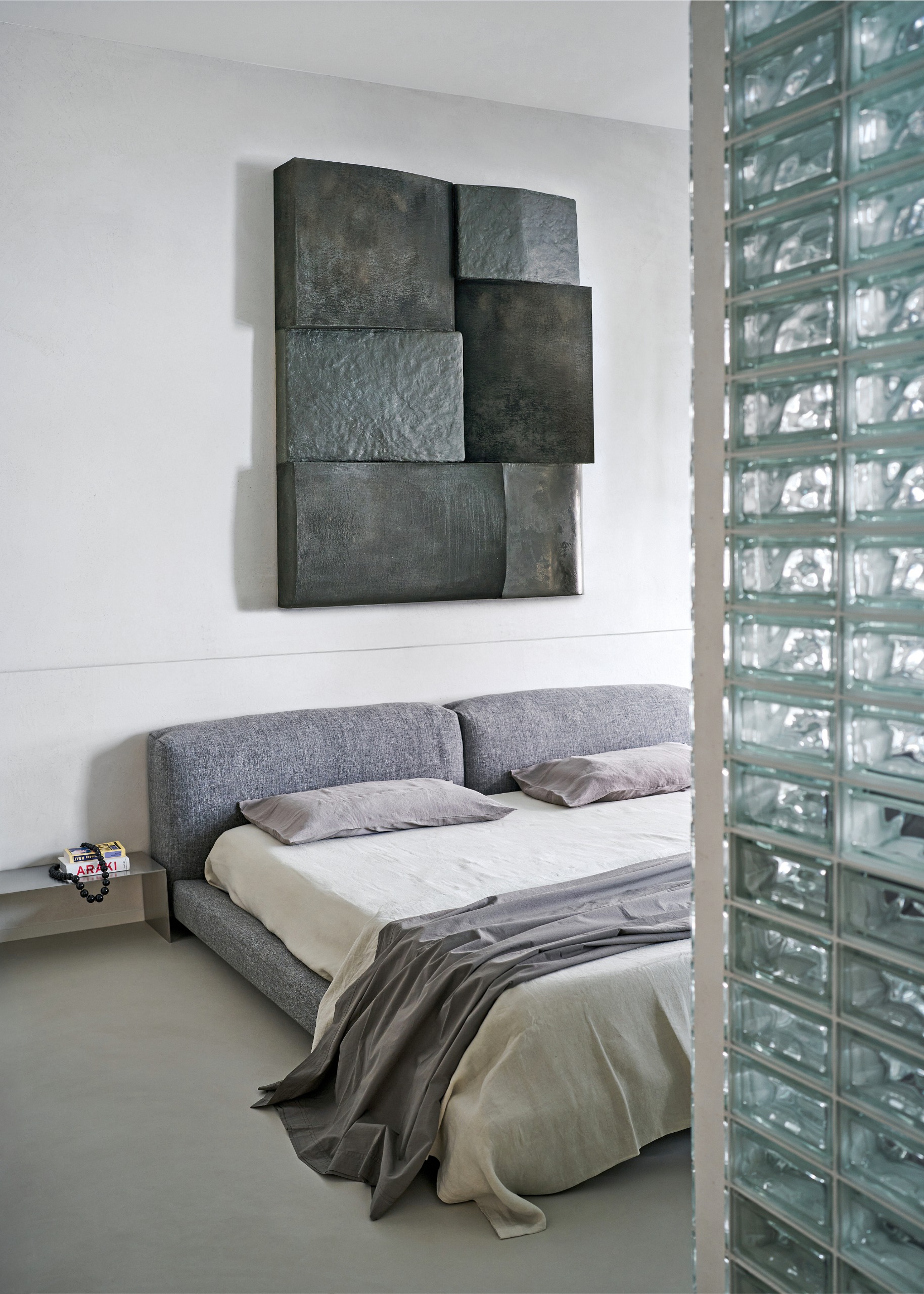 Bedroom with grey low-slung bed, grey tonal bedding, grey abstract artwork and glass block screen
