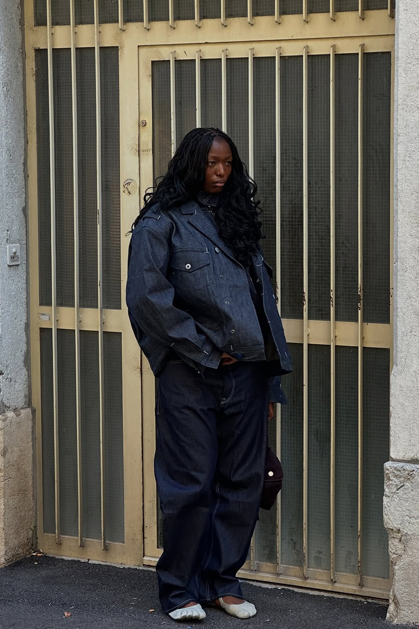 Berlin fashion trends are shown in a street-style photo of a woman standing outside in front of a door. She is wearing an oversized denim jacket over a denim top, styled with super-baggy dark-wash jean, white toe shoes, and a black shoulder bag