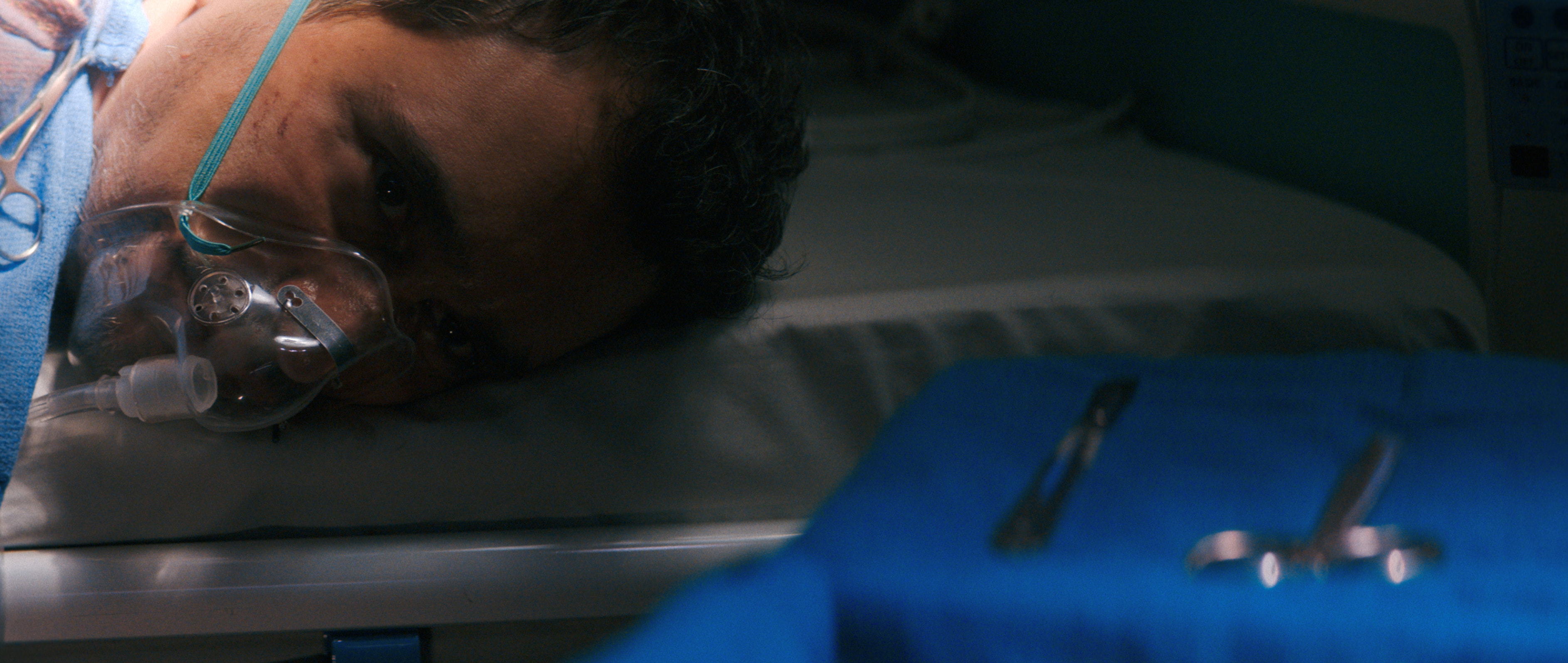 pluribus season 1 still of carlos manuel vesga as Manousos in an oxygen mask lying his head on an operation table with medical tools next to him