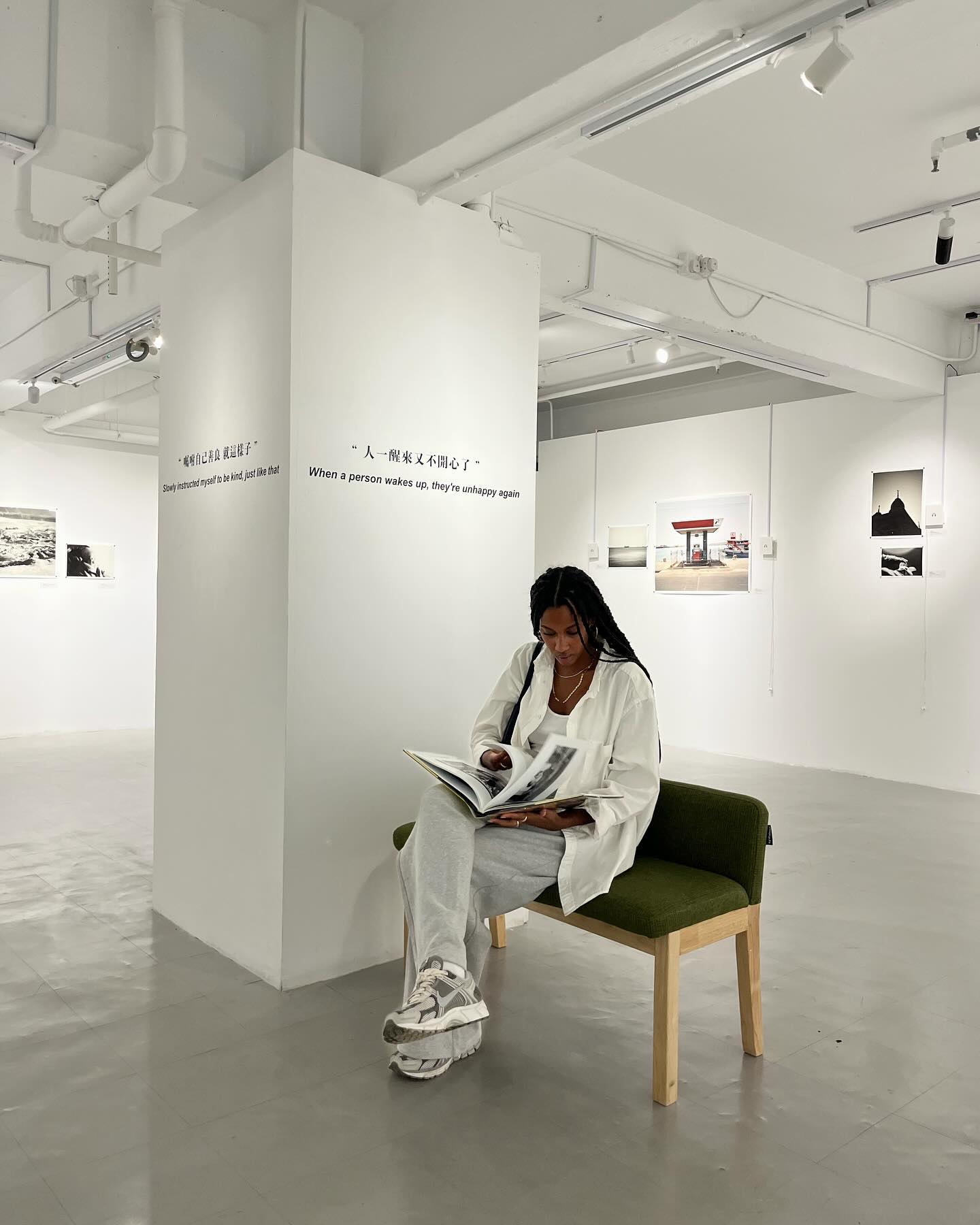 woman sitting on a bench in a museum gallery with white walls wearing white oversize button-down, gray sweatpants, and gray trainer sneakers