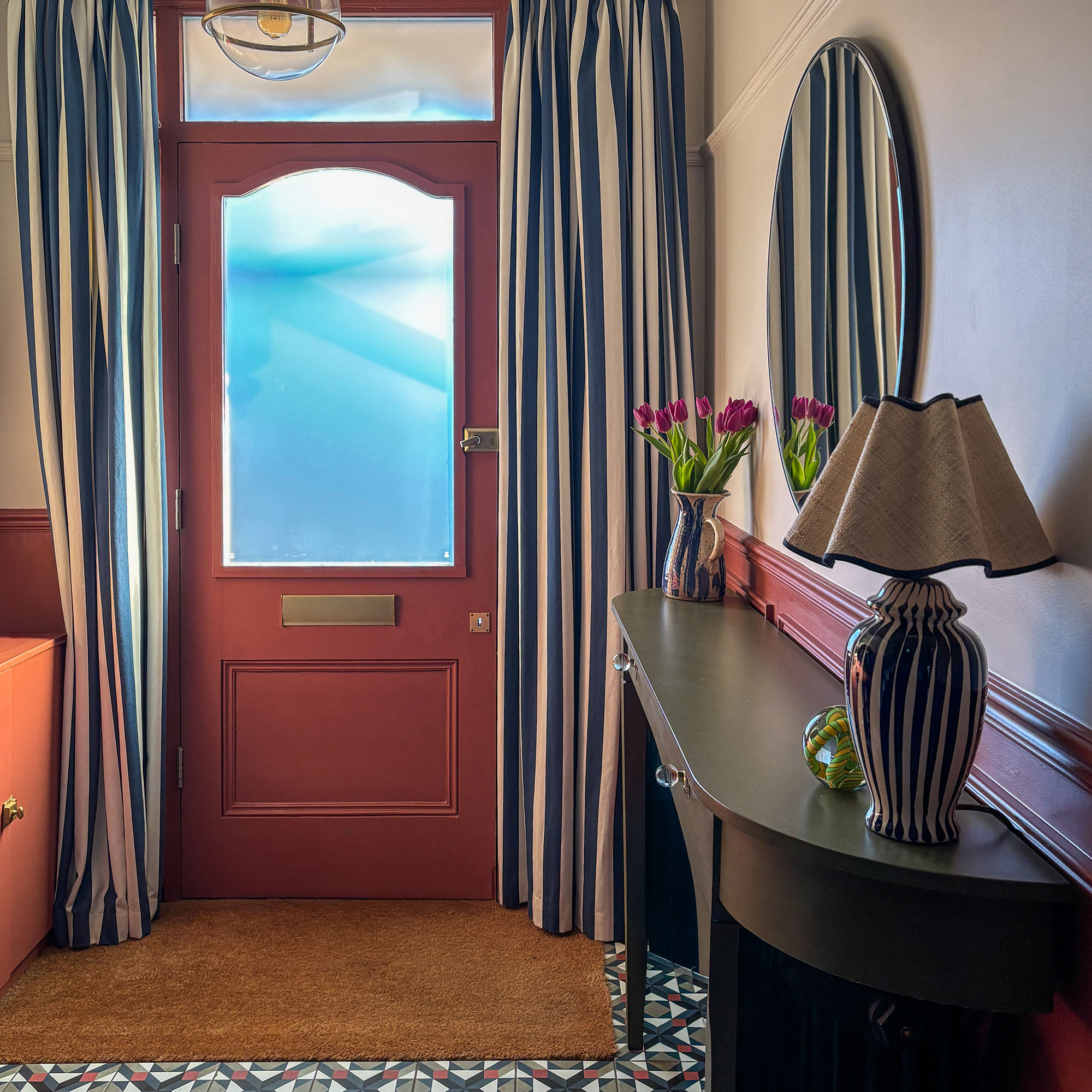 Hallway with terracotta front door, blue and white striped door curtains, dark coloured sideboard with vase and lamp topped with round mirror