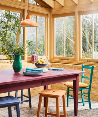 sunroom with yellow woodwork, red table, colored chairs, yellow pendant light