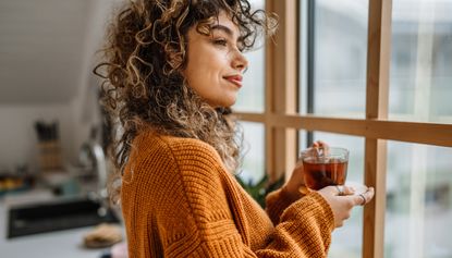 Young woman holding a cup of tea and standing by the window. Tea time rituals, morning rituals