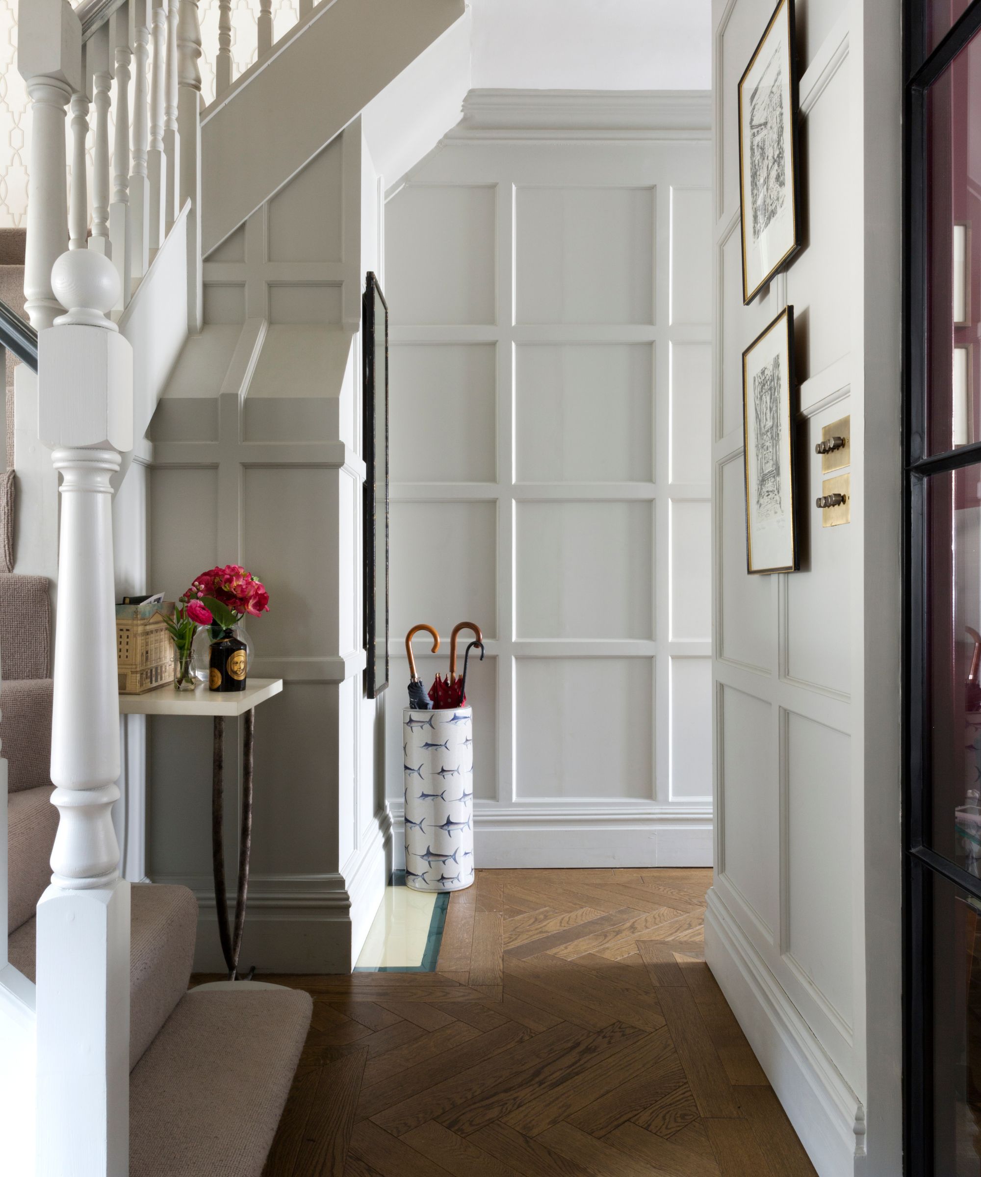 A bright entryway with white walls, wooden flooring, beige carpet running up the stairs, and a white umbrella stand patterned with fish holding three umbrellas. To the left is a small table with pink flowers on top.