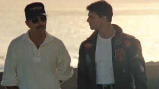 Tom Skerritt on the left in a hat and sunglasses talking to Tom Cruise in a white tee shirt and leather bomber jacket in Top Gun